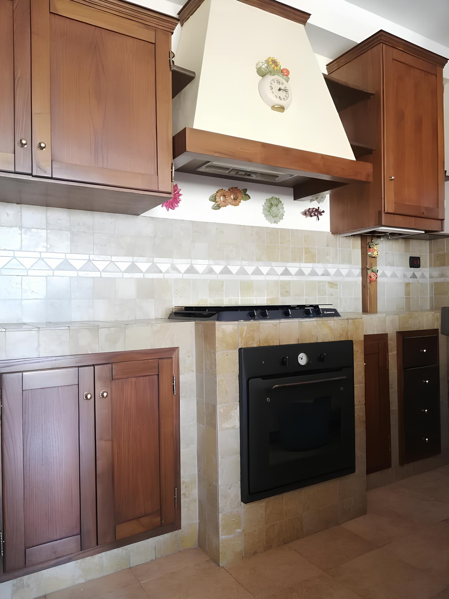 cucina Cucina con mobili in legno scuro, piano cottura nero integrato in muratura e cappa bianca, piastrelle bianche in fondo.