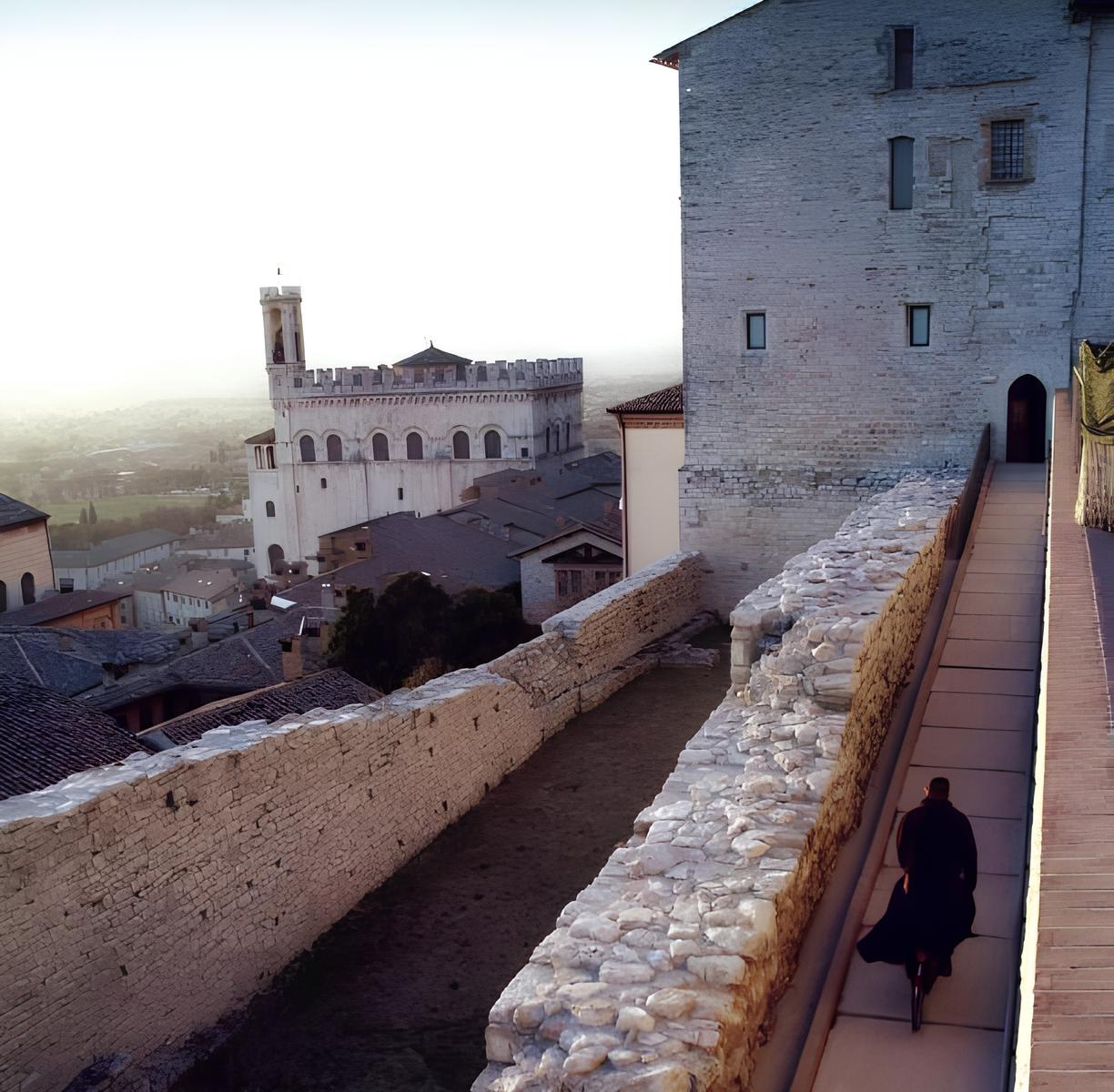 Veduta da una torre medievale con mura in pietra che si affacciano su un castello e la vallata sottostante al tramonto.